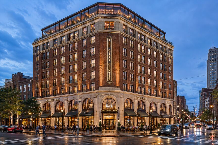Artist's conceptual rendering of The City Center — a refined brick corner building with a glassed rooftop restaurant and arched storefronts at dusk on a wet downtown street. Conceptual only; storefront signage illustrative.